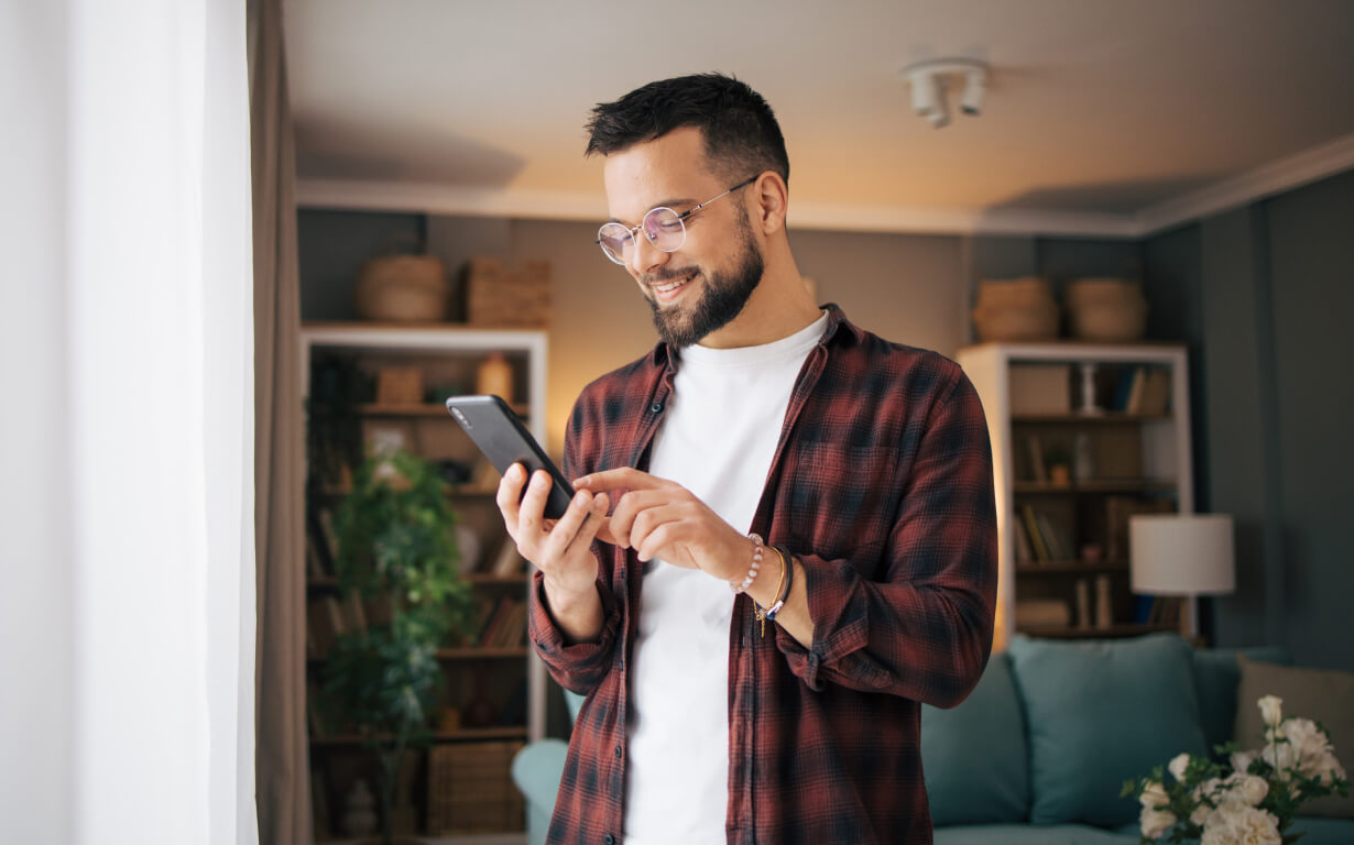 A man with a beard, wearing glasses and a plaid shirt, is standing indoors, smiling and using a smartphone. The background features shelves with baskets and a plant, and a softly lit room with a couch.