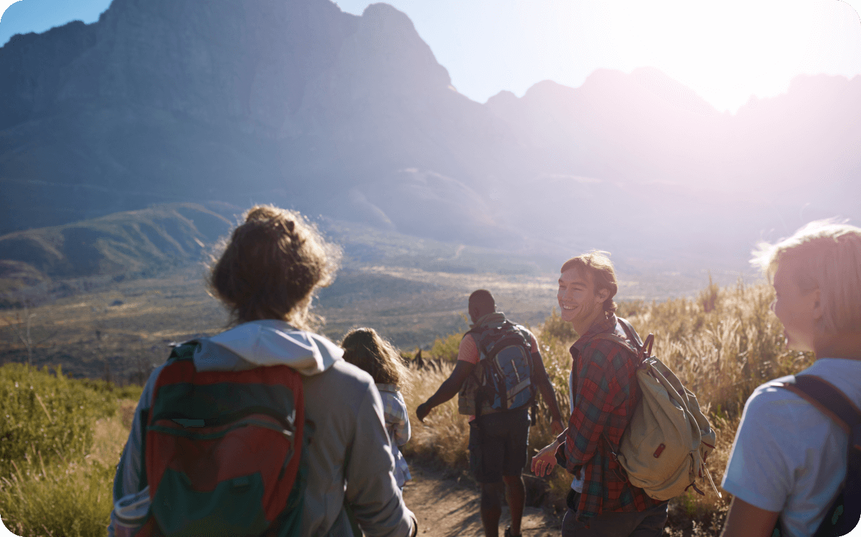 A group of hikers with backpacks walk along a trail with mountains in the background during their gap year abroad. One person is turning back and smiling at the camera. The sun is shining brightly, casting a soft glow over the scene.