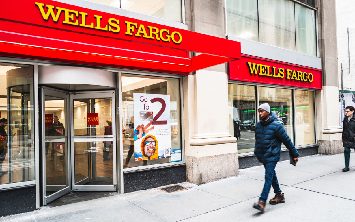 A Wells Fargo bank branch with red signage, recognized as one of the 5 Best US Banks, is situated on a city street. The entrance features glass doors and a promotional poster inside. Two pedestrians walk past the building on the sidewalk.