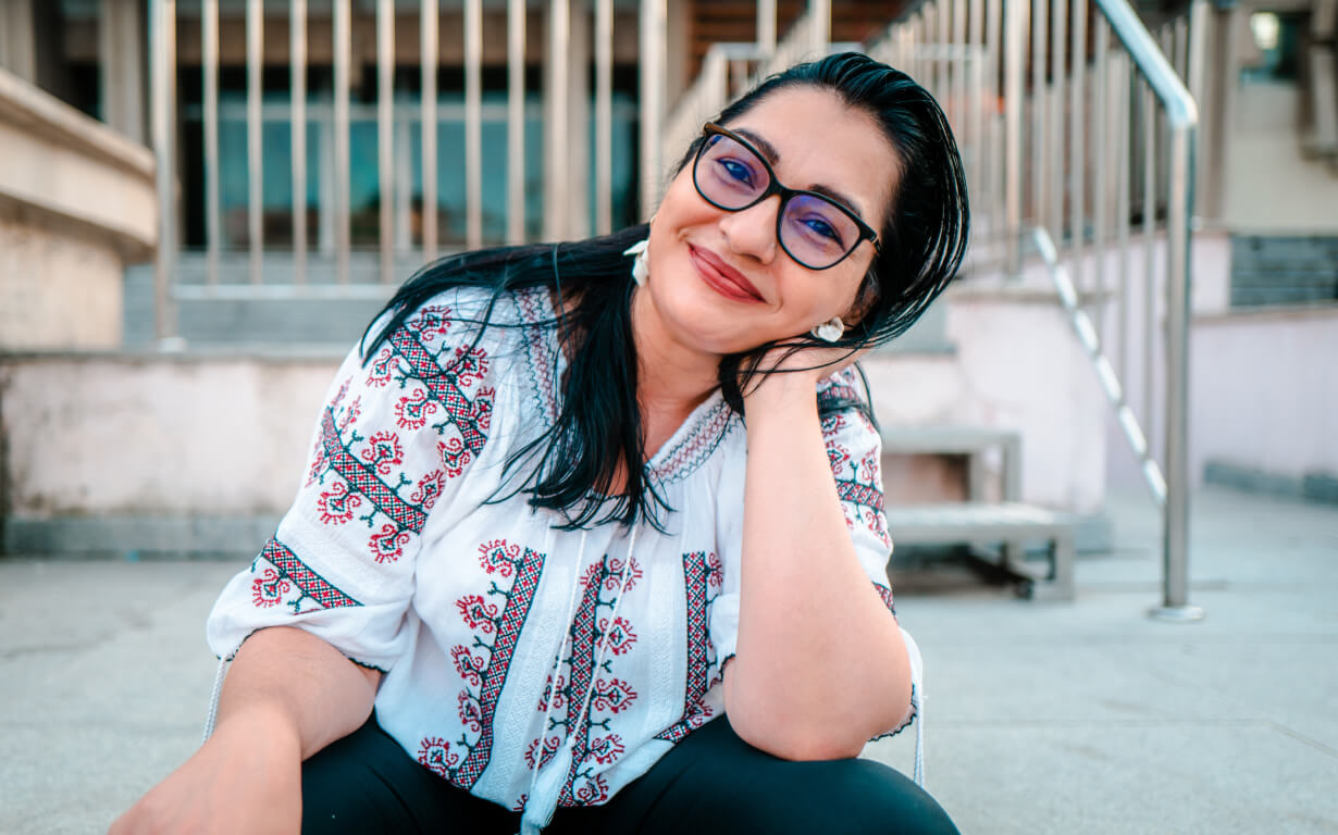 A woman wearing glasses and a white embroidered blouse, reminiscent of styles cherished in migrant communities, smiles while sitting outside. She rests her head on her hand, with stairs and metal railings in the background.