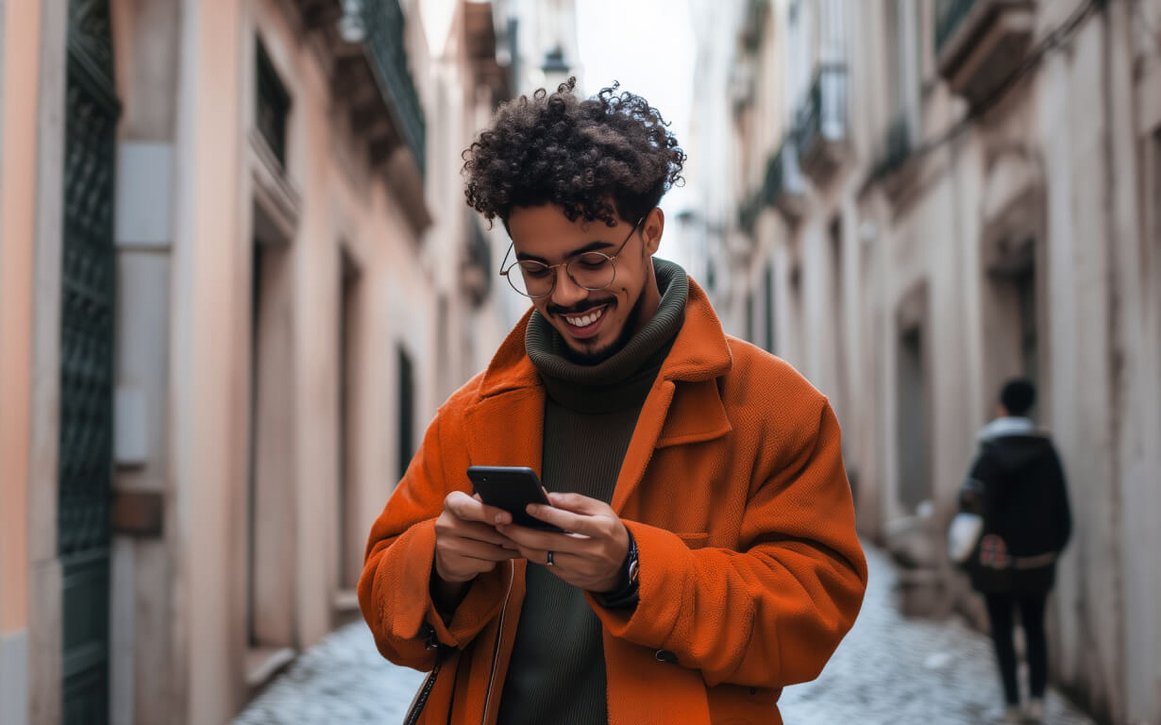 A man with curly hair and glasses is smiling at his smartphone, possibly checking how to receive money overseas in the US. Clad in an orange coat and green sweater, he strolls down a narrow, cobblestone street lined with historic buildings. Another person lingers in the background.