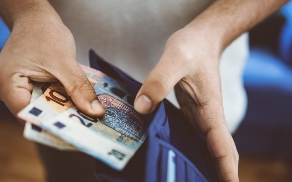 A person is holding a wallet and inserting Euro banknotes into it, perhaps preparing to send remittances. The denominations visible include 20 and 10 Euros. The person's hands are in focus while the background is blurred.