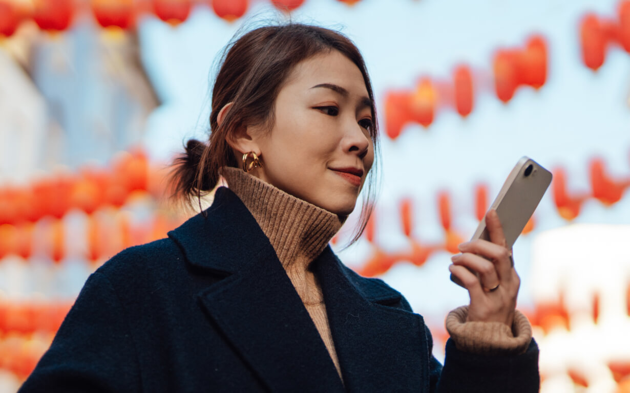 A woman in a dark coat and turtleneck sweater glances at her smartphone, her gentle smile reflecting the joy of staying connected. She stands outdoors amid rows of red lanterns that dance against the clear blue sky.