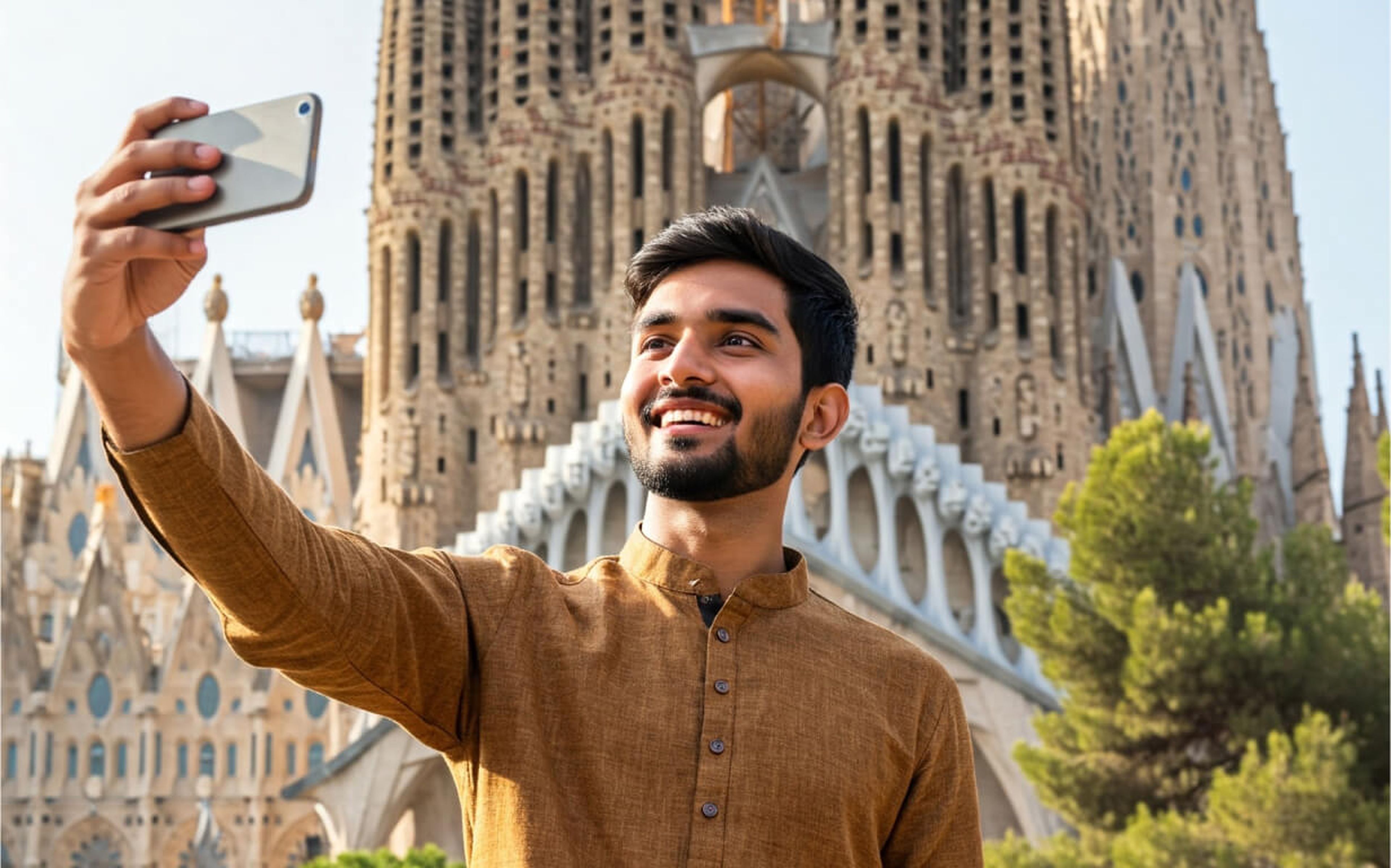 A young man in a brown shirt smiles while taking a selfie in front of the ornate facade of the Sagrada Familia basilica in Barcelona, Spain, capturing his excitement about moving to Spain on a sunny day.