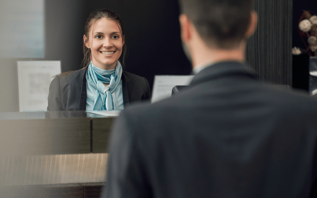 A smiling woman in professional attire and a scarf stands behind a reception desk, facing a man in a suit who has his back to the camera. The setting appears to be the reception area of one of the best European banks or possibly an upscale hotel.