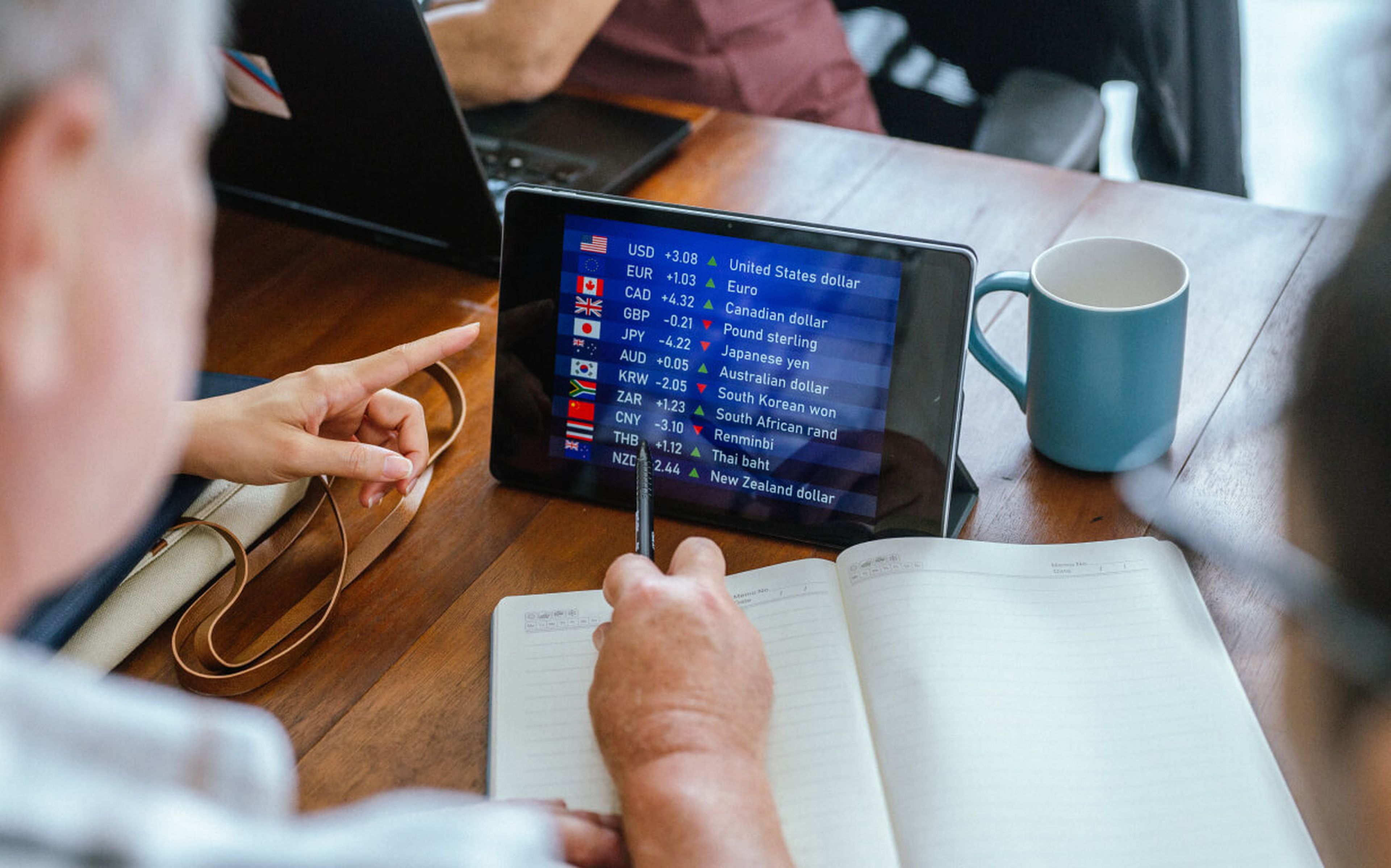 Two people reviewing fluctuating exchange rates on a tablet. The screen displays flags, currency codes, and detailed charts. One person points at the display with a pen. A notebook and a steaming mug sit on the wooden table, creating an ideal setting for their financial analysis.