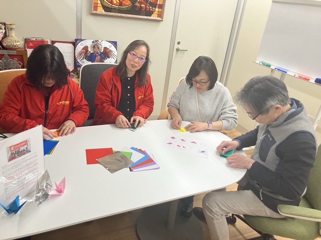 Four adults sit around a white table folding colorful origami paper. Two women in red jackets are on the left, while two people on the right focus on their paper. Origami cranes and instructions are on the table.