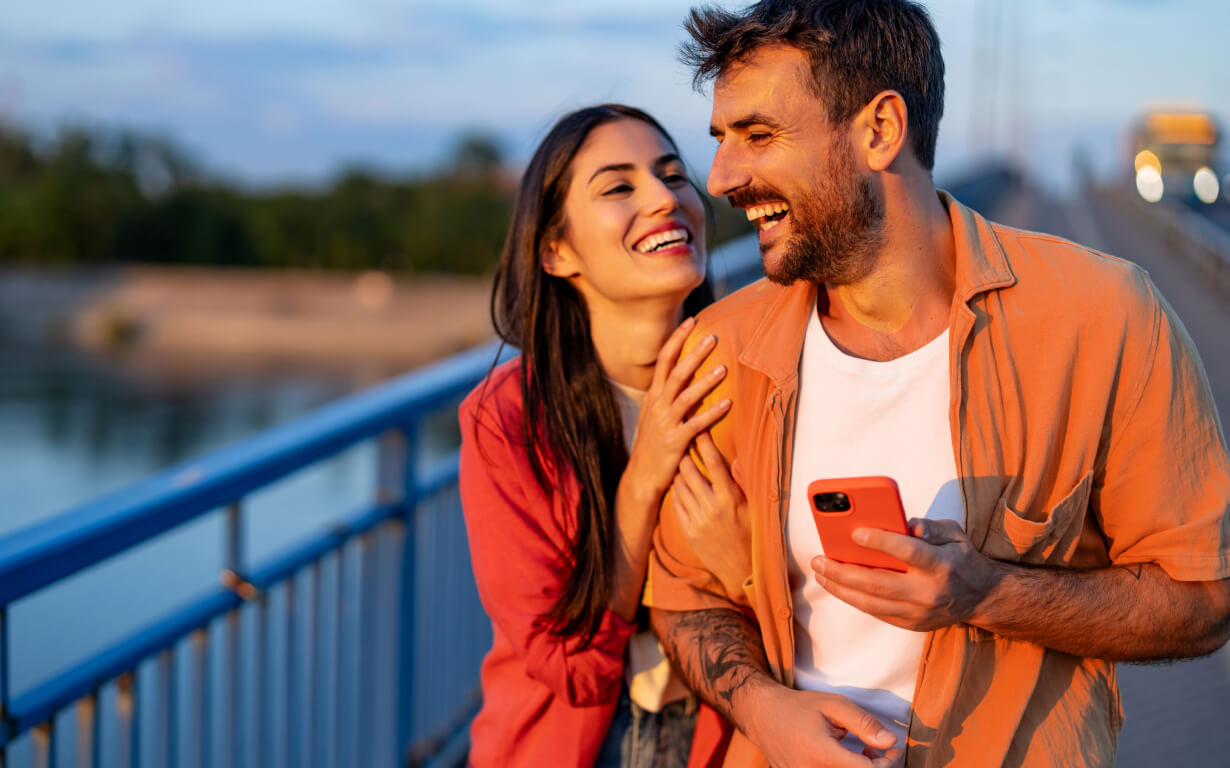 A happy couple stands on a bridge at sunset, capturing memories with their smartphone. The man, in an orange shirt, and the woman in red share a smile as they lean into each other. As the river softly flows behind them, they're reminded of modern conveniences.