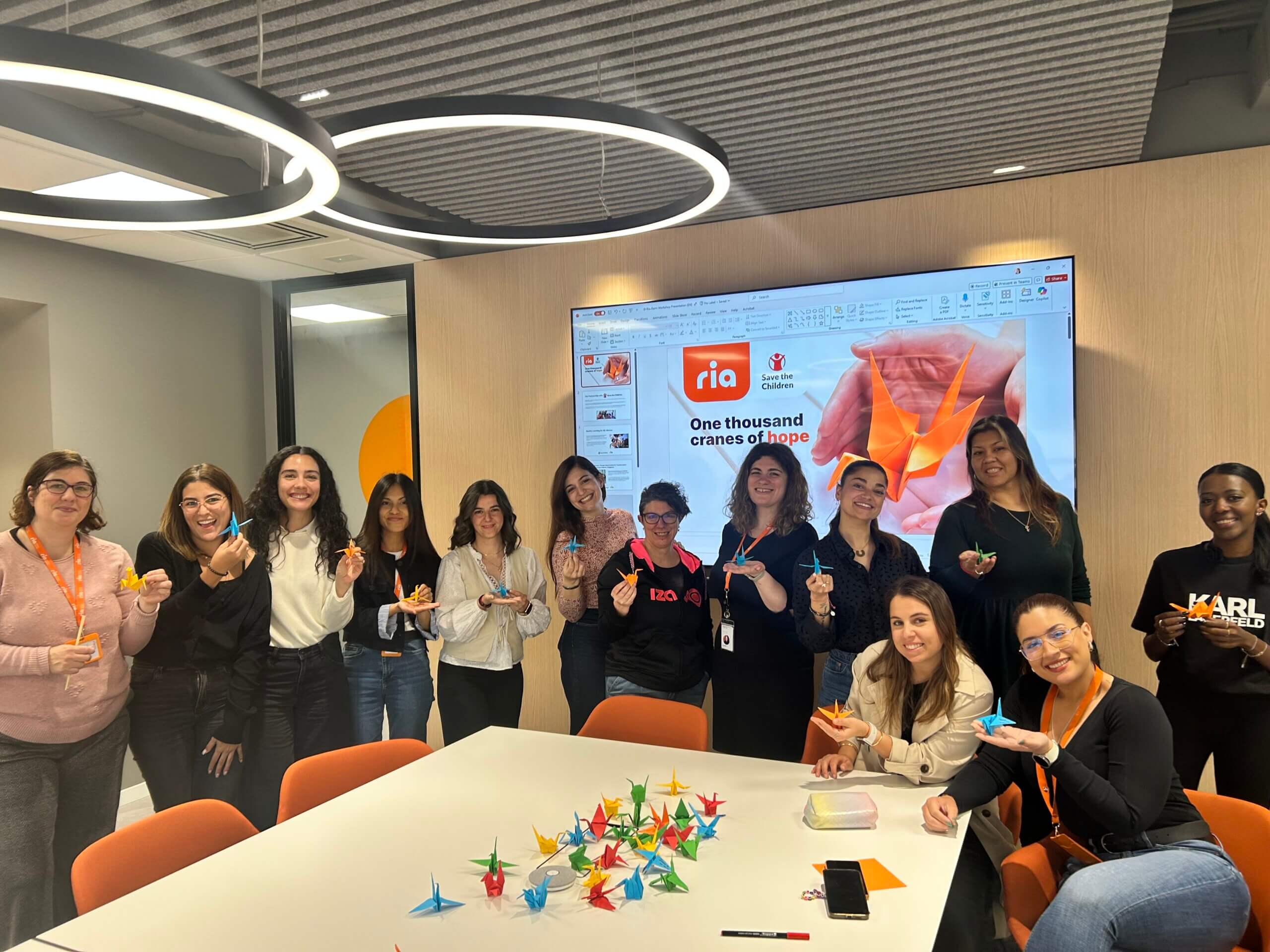 A group of people in an office stand and sit around a table, smiling and holding colorful origami cranes. On the table are many more cranes. A large screen behind them displays “One thousand cranes of hope” with an orange origami crane.