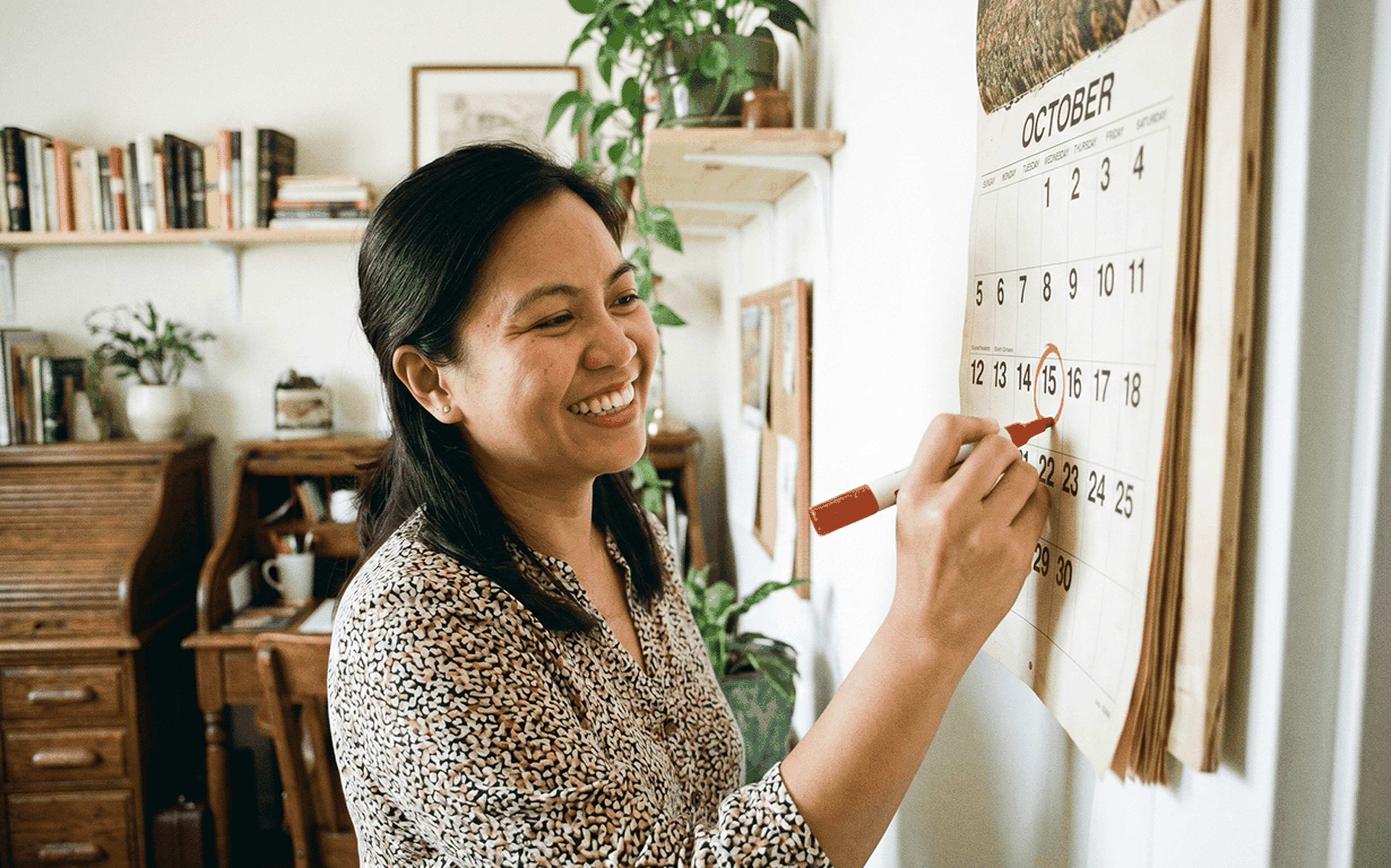 A woman smiles while circling October 16th on a wall calendar with a red marker, likely marking her money sending routine, in a cozy, plant-filled room with bookshelves and a wooden desk in the background.