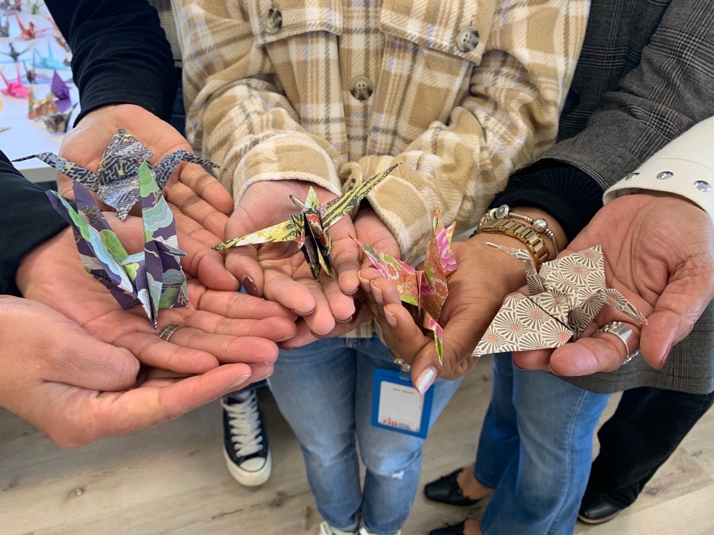 Four people standing in a group hold colorful origami paper cranes in their outstretched hands. They wear casual clothing, including plaid and blazers, and name badges are partially visible.
