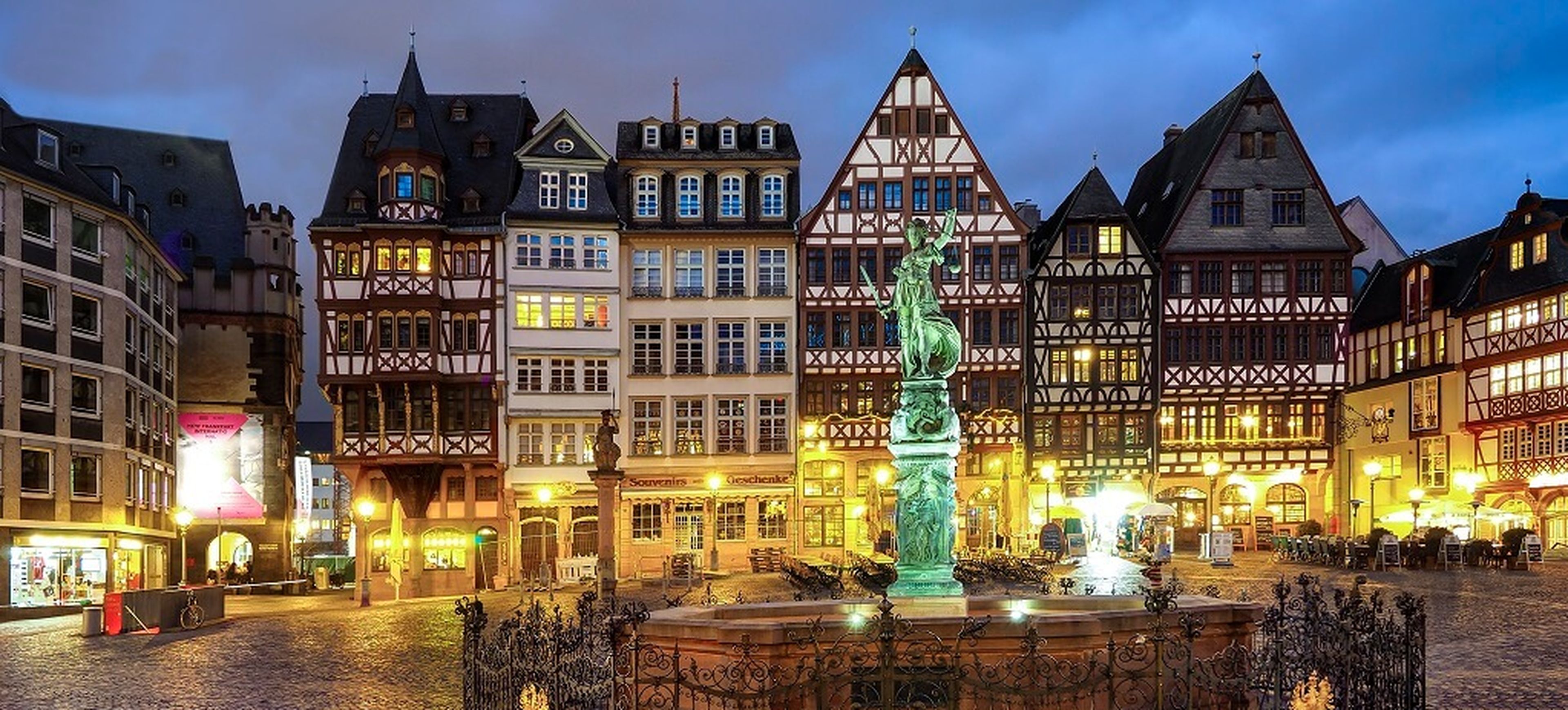 The image shows a historic European town square at dusk, featuring half-timbered buildings with illuminated windows. A central statue with a fountain is in the foreground, surrounded by streetlights and outdoor seating. The sky is a gradient of deep blue.