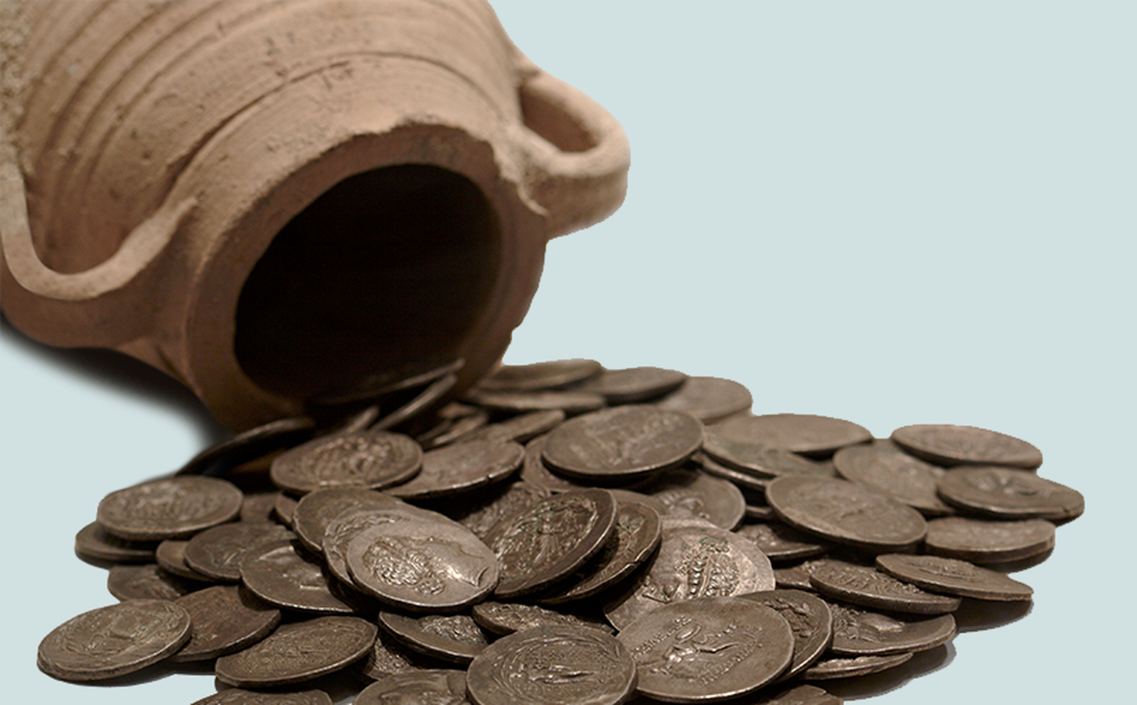 A sideways clay jar with two handles spilling out numerous ancient coins onto a surface. The coins are dark and feature engravings. The background is light, accentuating the jar and coins.