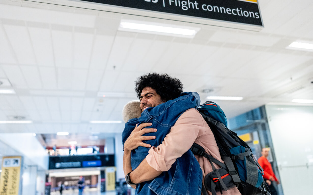 In an airport terminal bustling with travelers, two people embrace joyfully, one sporting a backpack. Above them, a sign for flight connections looms as the backdrop teems with anticipation and Valentine's Day warmth lingers in the air.