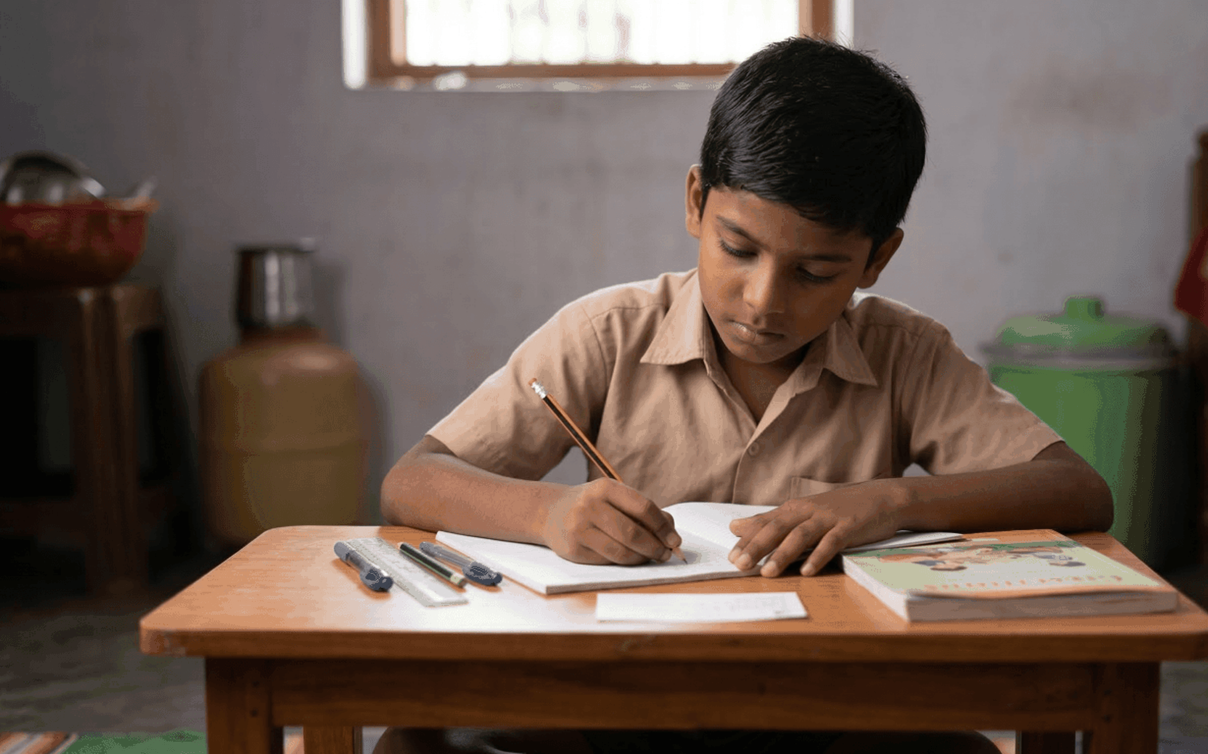 A young boy sits at a wooden desk, focused on writing in a notebook with a pencil. Several pens, a book, and notes about remittances are on the desk. The room has simple furnishings and daylight comes through a window behind him.