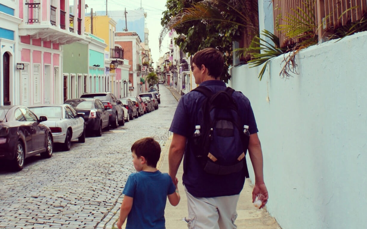 A man and a child walk hand in hand down a cobblestone street lined with parked cars and colorful buildings, as they embrace the adventure. The man carries a backpack with water bottles, and the street leads uphill, bordered by a white wall and plants.