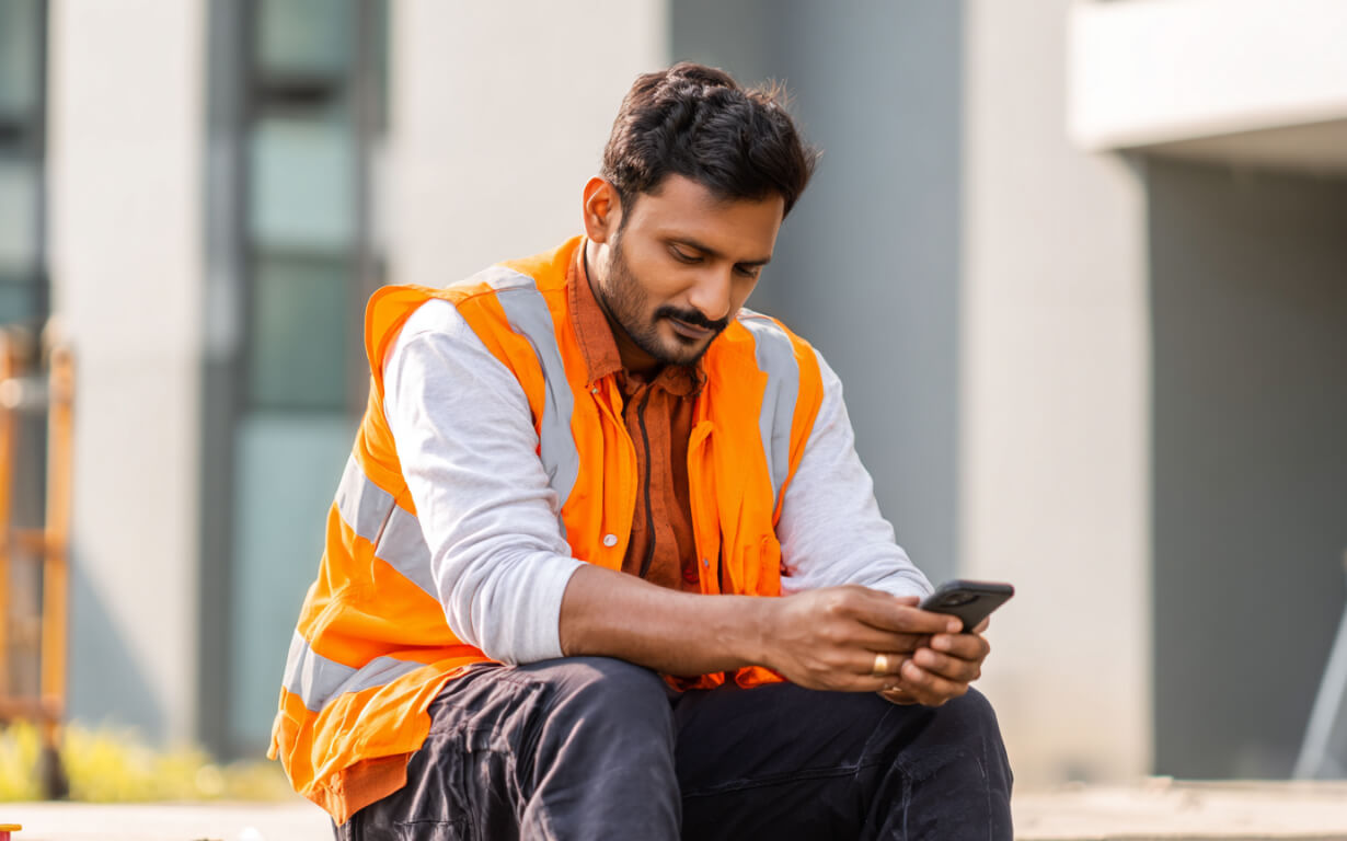 A construction worker wearing an orange safety vest sits outdoors, looking at his smartphone as he prepares to Send Money to India.