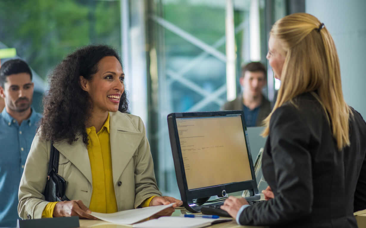 A woman in a yellow coat is smiling as she discusses the safest ways to send money with a receptionist behind a desk with a computer screen. A man stands in line behind her, all within the sleek setting of a modern office or hotel lobby framed by glass walls.