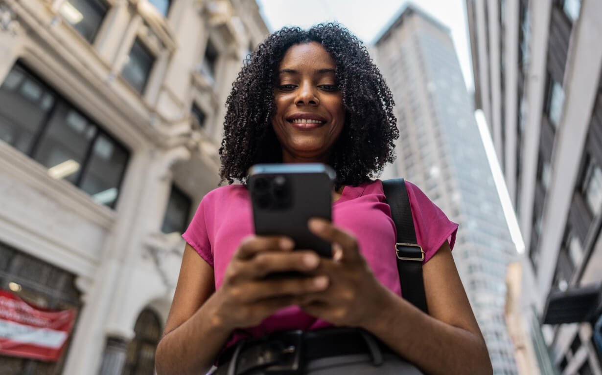 A woman with curly hair, wearing a pink top, smiles as she looks at her smartphone. She is standing on a city street, with tall buildings in the background. The shot is taken from a low angle, emphasizing the architecture around her as she prepares to send remittances back home.