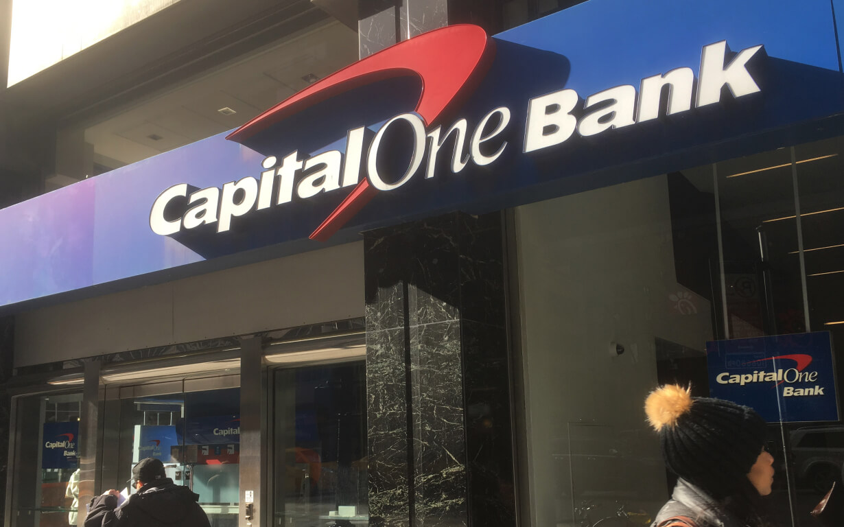 A Capital One Bank branch, one of the 5 Best US Banks, features a blue sign with white lettering on its exterior. People in winter clothing stroll by on the sidewalk.
