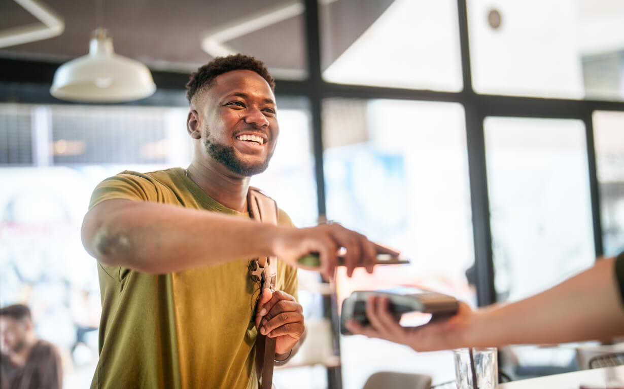 A smiling man in a green shirt is using his digital wallet to make a contactless payment with his smartphone at a cafe. He holds his phone near the payment terminal held by another person, ensuring a smooth transaction.