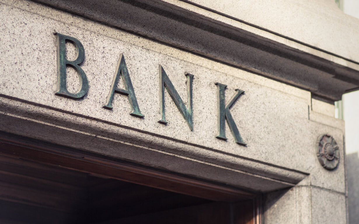 Facade of a stone building with large, metal letters spelling "BANK" above a doorway. The architectural details suggest a classic or historic style.