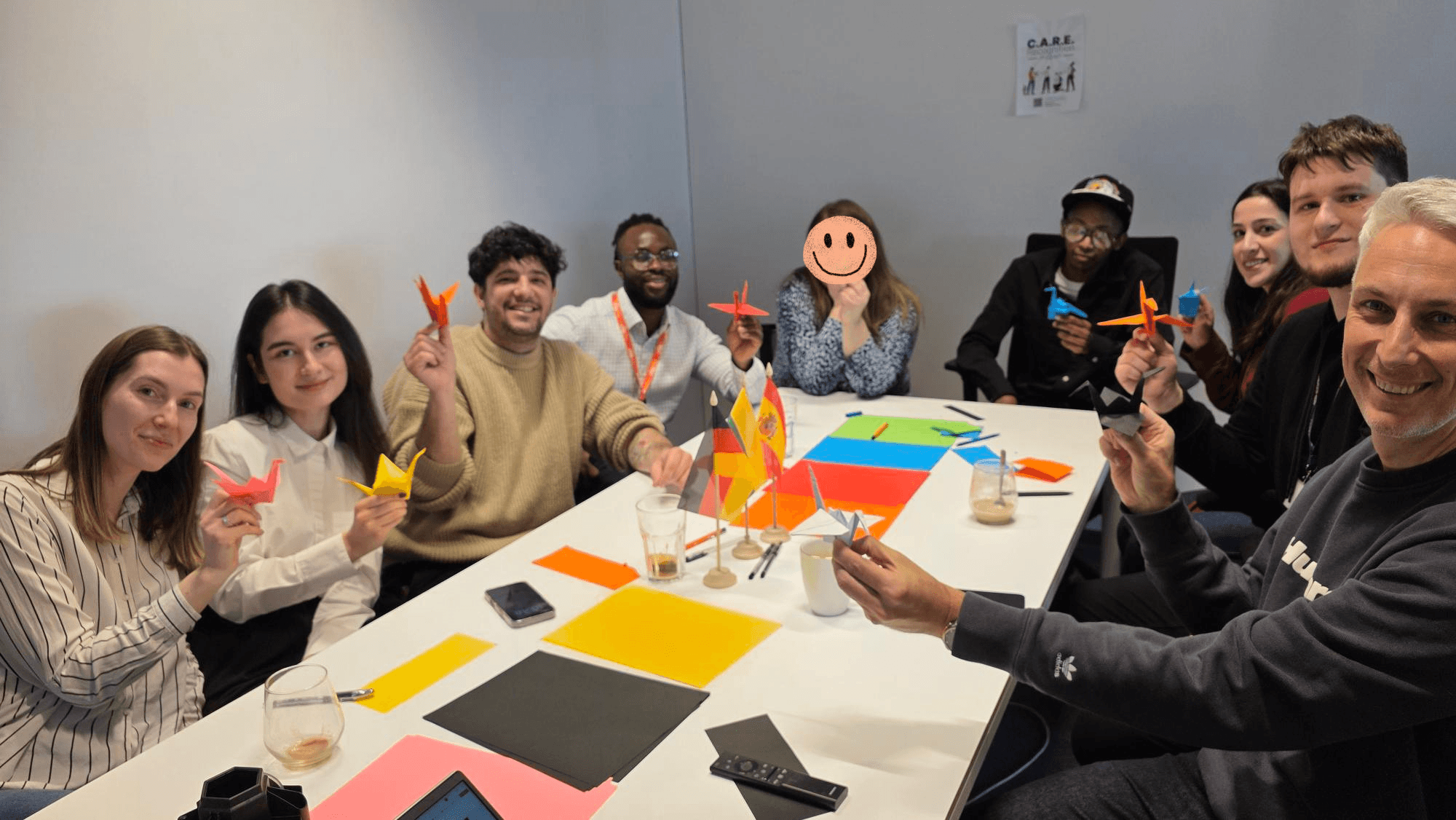 A group of people sits around a table holding colorful origami cranes, smiling at the camera. The table is covered with colored paper and drinks. One person's face is covered with a smiley face drawing.