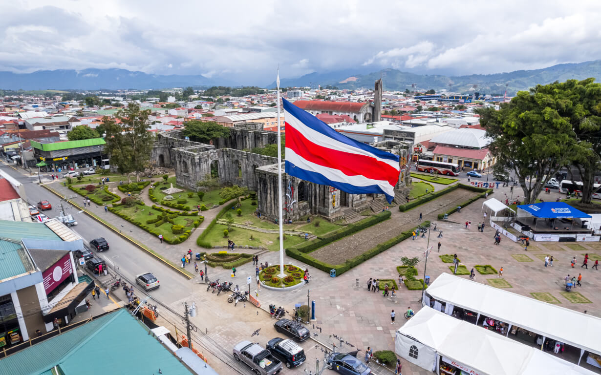 A large Costa Rican flag waves over a city square with the ruins of a historic building at the center. People walk around the landscaped area, which is surrounded by streets, vehicles, and buildings. Mountains and clouds are visible in the background.