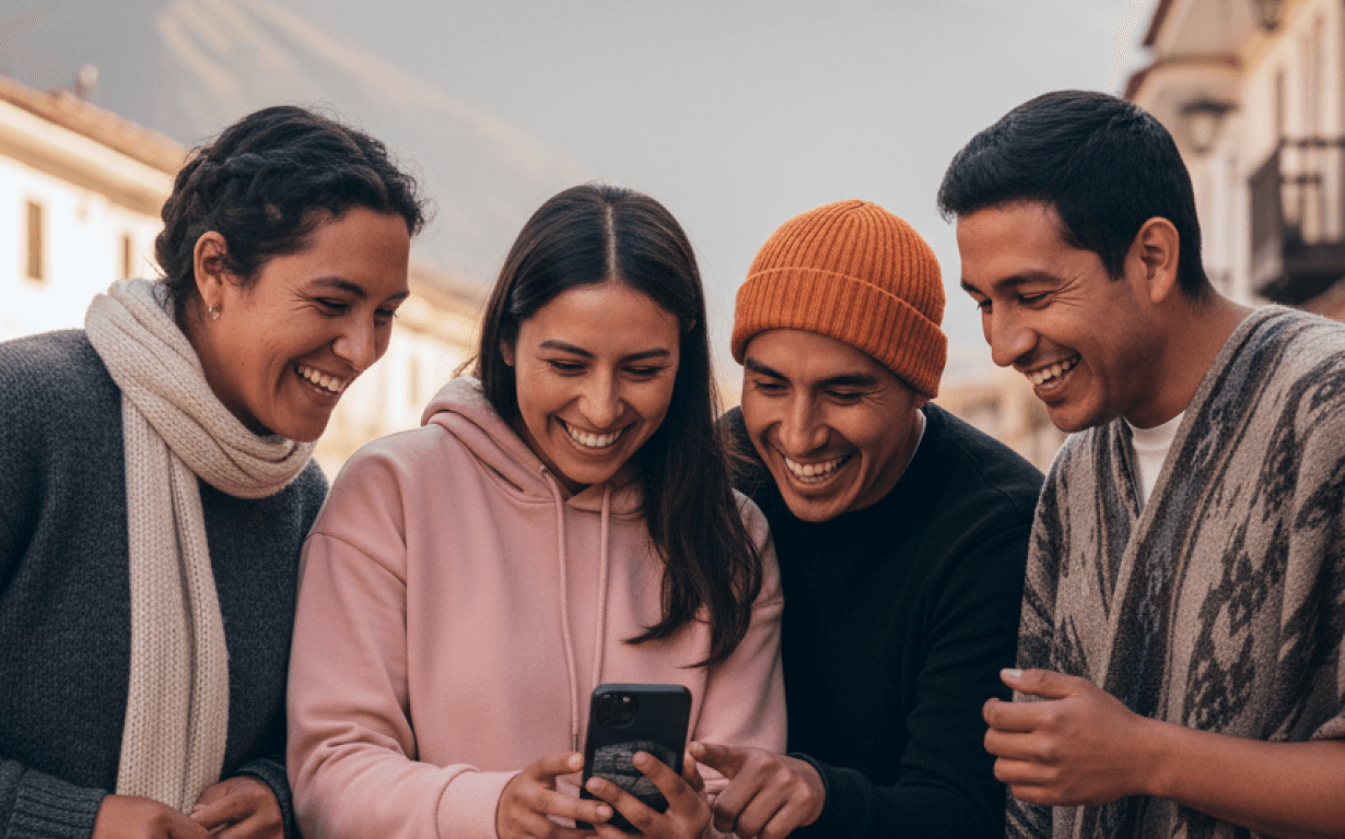 Four people stand outdoors smiling and laughing as they look at a smartphone, perhaps discovering an easy way to send money to Peru. Dressed warmly in sweaters and a beanie, they appear happy and engaged with buildings in the background.