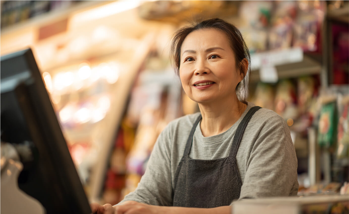 A woman wearing a gray shirt and dark apron smiles while standing behind a cash register in a brightly lit store, where customers can Send Money Without a Bank Account. Shelves of products are visible in the background.