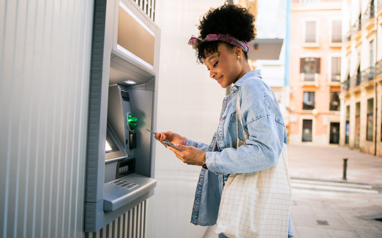 A person in a denim jacket stands at an outdoor ATM, holding a bank card. They're looking at their phone, possibly checking exchange rates or transaction details. The background features colorful buildings under a clear sky.