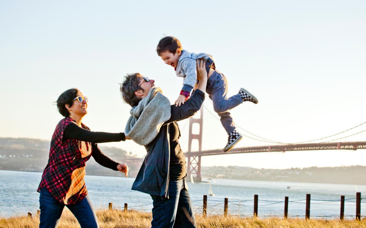 A family of three enjoys a playful moment outdoors near the water, with a bridge in the background resembling those iconic scenes. One adult throws child into the air, while the other stands nearby, smiling under a clear, sunny sky.