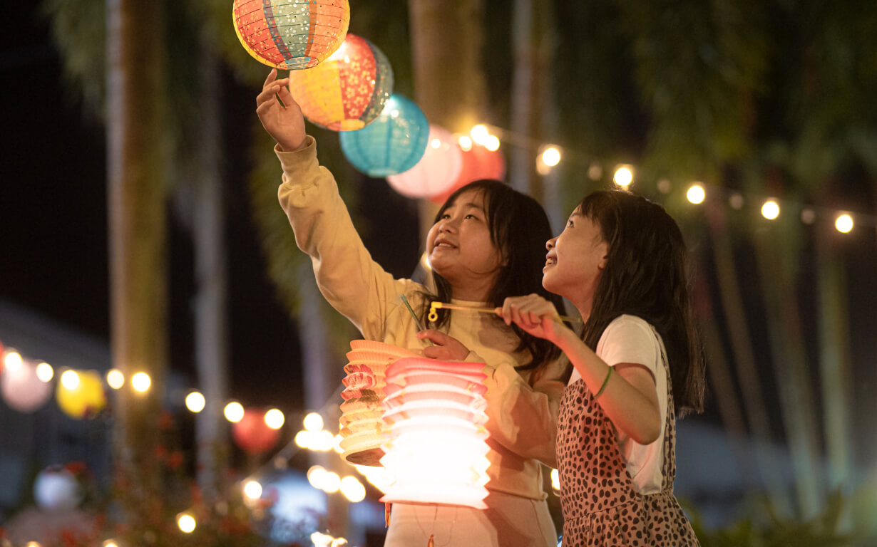 Two children smile and hold colorful lanterns at a nighttime outdoor festival celebrating the Lunar New Year. String lights and blurred foliage create a festive atmosphere.
