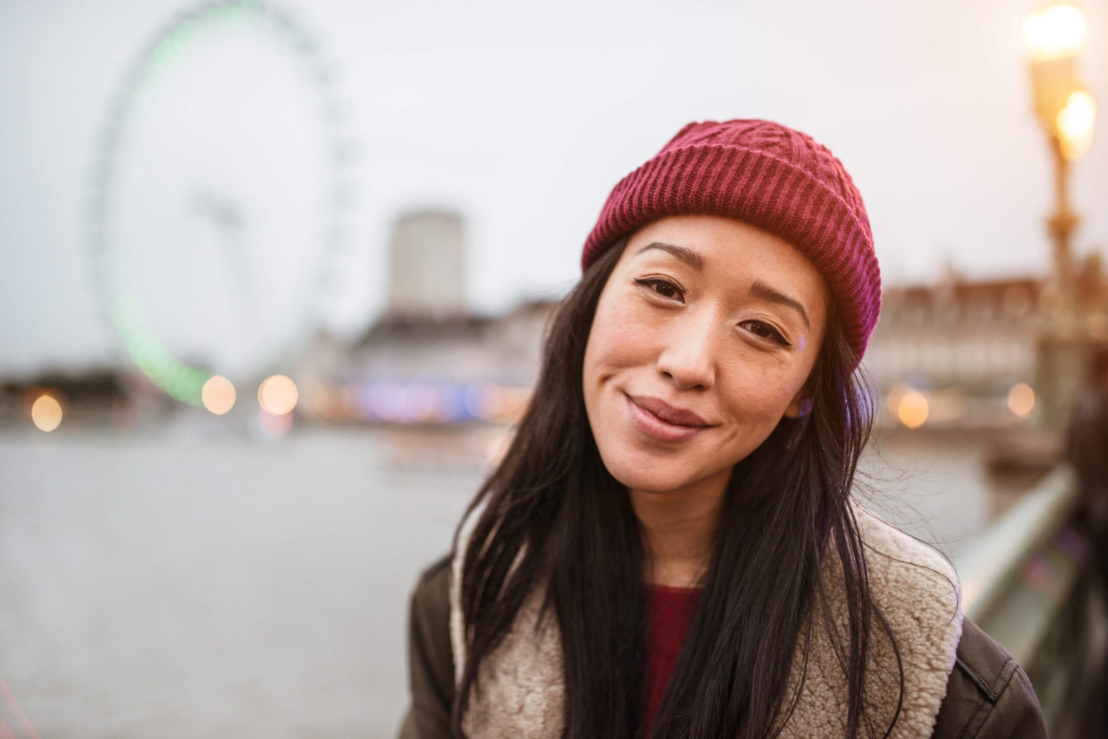 A woman wearing a red beanie and a fur-lined jacket smiles at the camera. The background, with its large ferris wheel, blurred city buildings, and river, hints at an urban setting enriched by vibrant migrant communities. The lighting suggests a cloudy or overcast day.