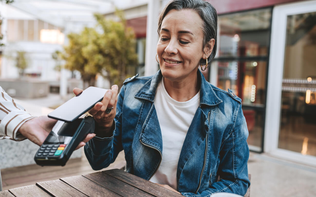 A person in a denim jacket smiles while holding a smartphone near a payment terminal, likely using their digital wallet for a contactless payment, at an outdoor café.