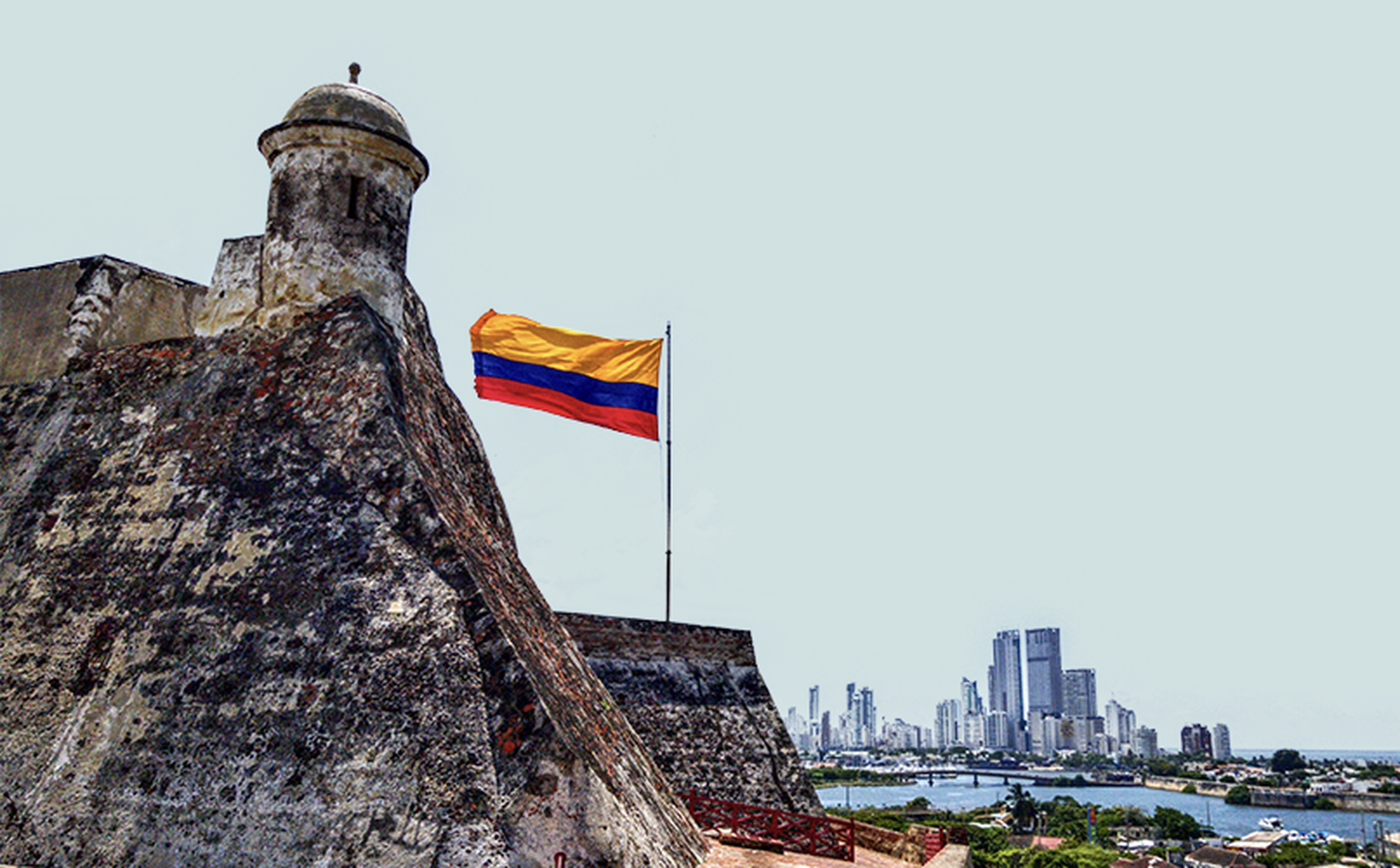 A large stone fortress with an ancient watchtower and a waving Colombian flag is positioned on a hilltop. In the background, there is a modern city skyline, reflecting the transformations brought by remittances and migration in Colombia, with tall buildings and water bodies under a clear sky.