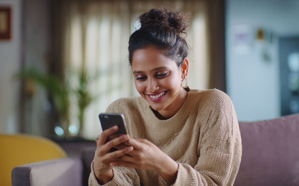 A woman with curly hair in a bun, wearing a beige sweater, sits on a couch smiling while looking at her smartphone, perhaps about to Send Money to India. A window with curtains and indoor plants are softly visible in the background.