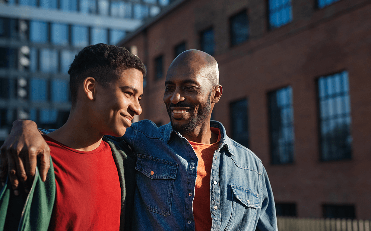 A smiling young man and an older man share a moment outdoors, discussing international money transfers. With the older man's arm around the younger's shoulders, they stand in front of urban buildings, both looking content and wearing casual clothing.