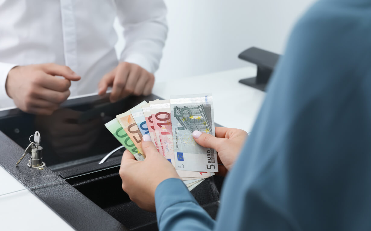 A person in a blue shirt holds several euro banknotes at a bank counter, while another in a white shirt stands across the desk.