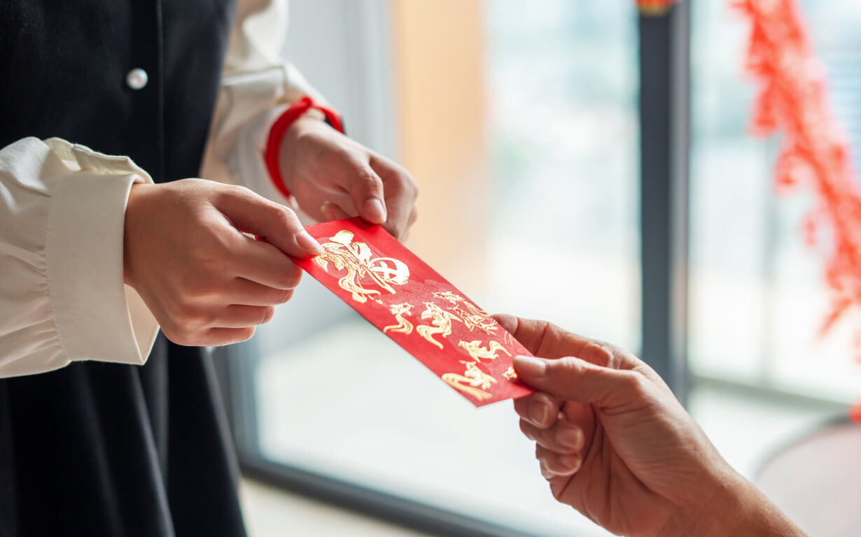 A person hands a red envelope adorned with gold designs to another, capturing the festive spirit of Lunar New Year. Indoors, the softly blurred background reveals glimpses of a window and vibrant red decorations.