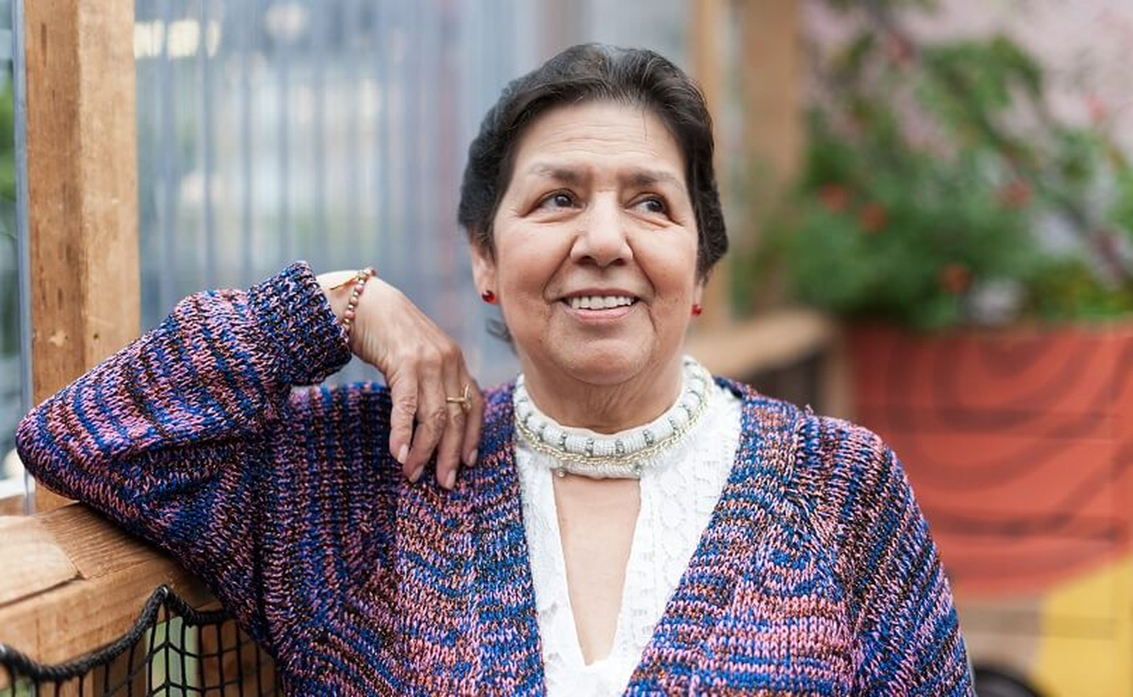 An elderly woman with short dark hair smiles while sitting outdoors. She is wearing a multicolored knitted sweater over a white blouse and resting her left arm on a wooden railing. The background features a colorful mural and potted plants.