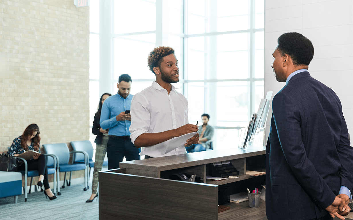 A man stands at a reception desk discussing international money transfers with the receptionist in an office lobby. In the brightly lit space, people are sitting and standing in the background, some using their phones to check tax updates. Large windows flood the room with natural light.