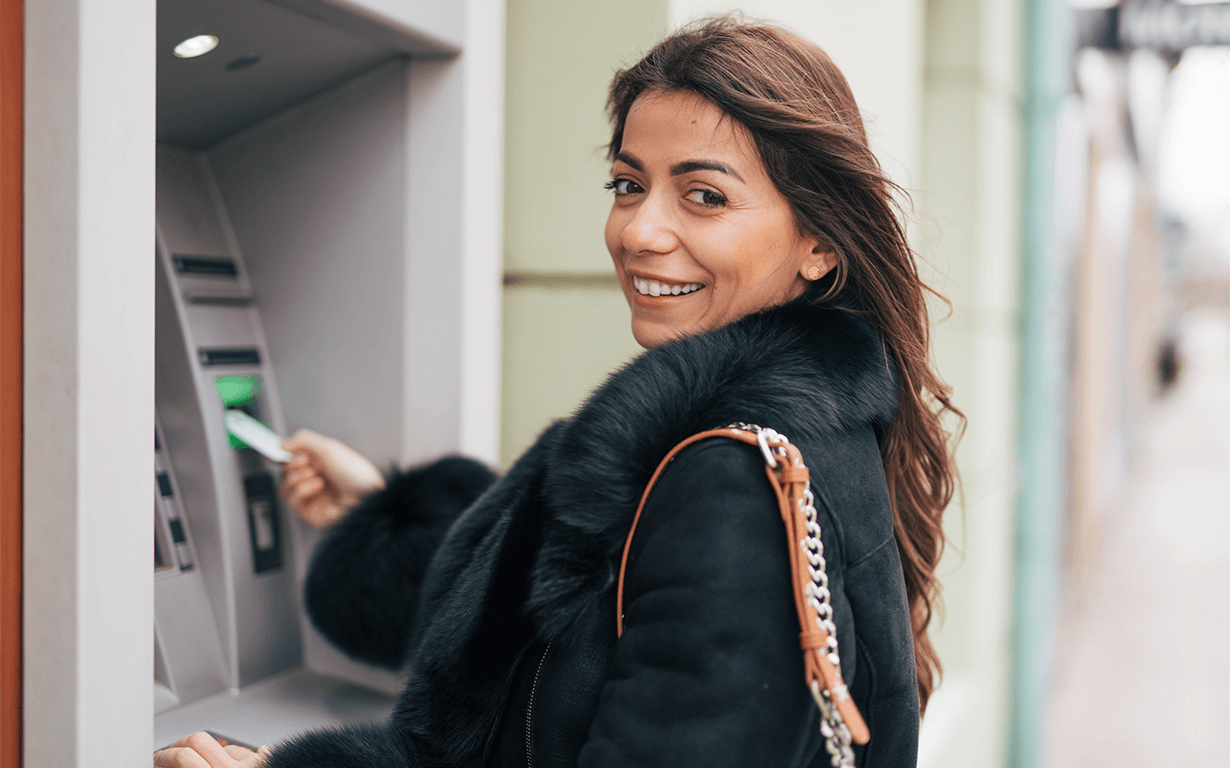 A person in a black fur-lined coat is using an ATM for international money transfers outdoors. They're smiling and looking over their shoulder at the camera. A beige building and a sidewalk are in the background, perhaps savoring a small victory amid tax season errands.