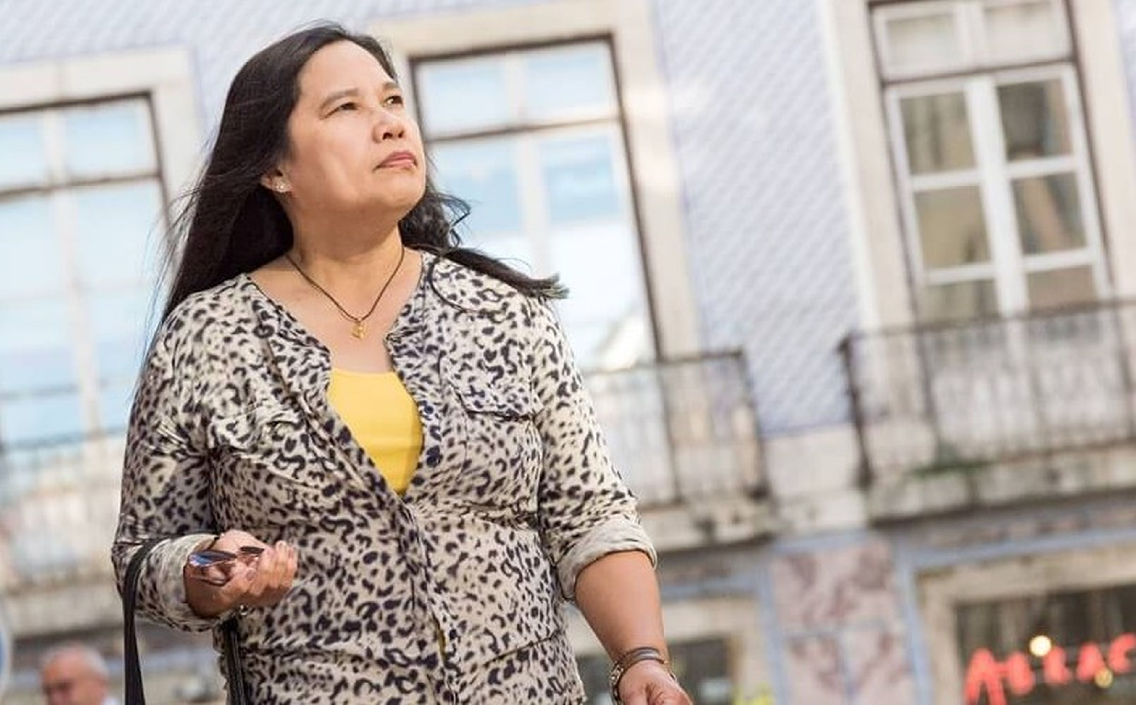 A mother from the Philippines with long dark hair, wearing a leopard print blouse over a yellow top, walks outside holding a cell phone. She looks upwards with a serious expression. Behind her, there are buildings with large windows and balconies.