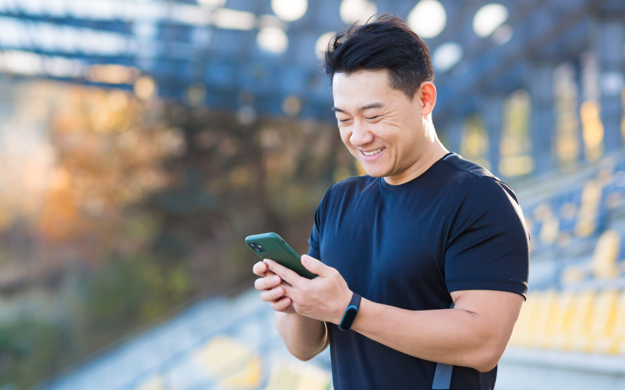 A man in a black T-shirt is smiling while using WeChat on his smartphone. He is standing in an outdoor setting with rows of yellow seats and a modern, open structure in the background.