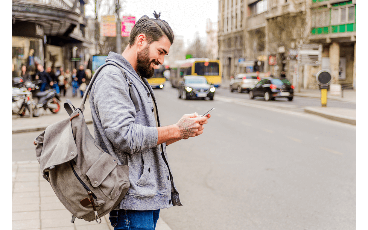 A bearded man with a gray backpack and a casual gray hoodie stands on a city sidewalk, smiling as he looks at his phone. Vehicles and buildings, including branches of some of the best European banks, are visible in the background, along with other pedestrians.