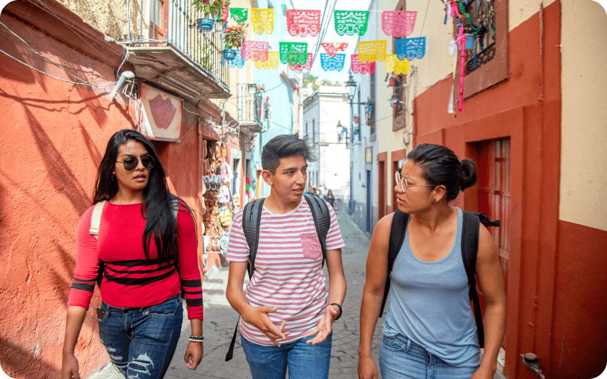 Three people walk down a colorful, narrow street adorned with vibrant paper cutouts. The street is lined with red and orange buildings, one of which has souvenirs displayed outside. They appear engaged in conversation about moving to Mexico and wear casual clothing.