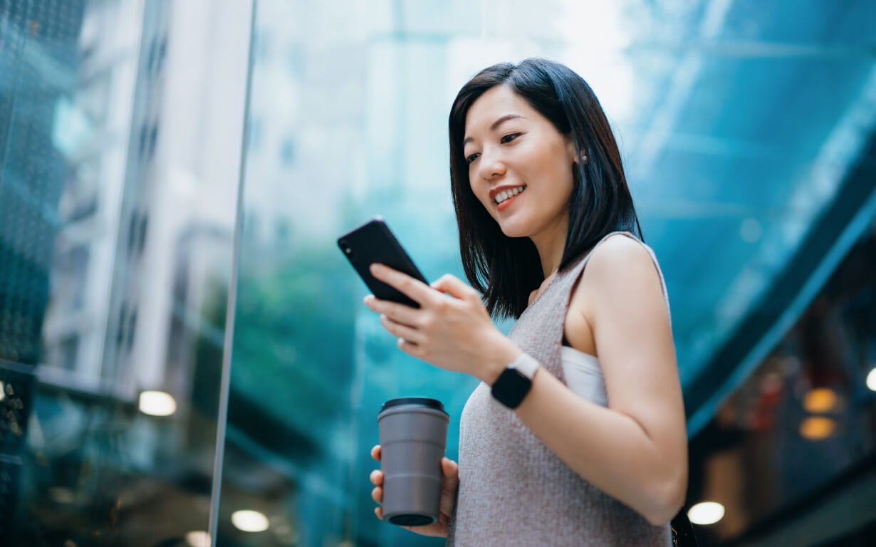 A woman stands outdoors in a city, smiling. She holds a coffee cup and wears a smartwatch, with modern buildings and a glass structure gleaming in the background.