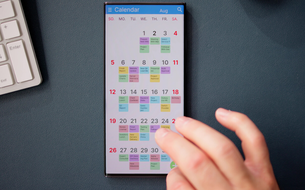 A person interacts with a smartphone displaying a digital calendar for August, filled with colored events, including currency exchange reminders. A glimpse of a computer keyboard is visible in the upper left corner of the image.