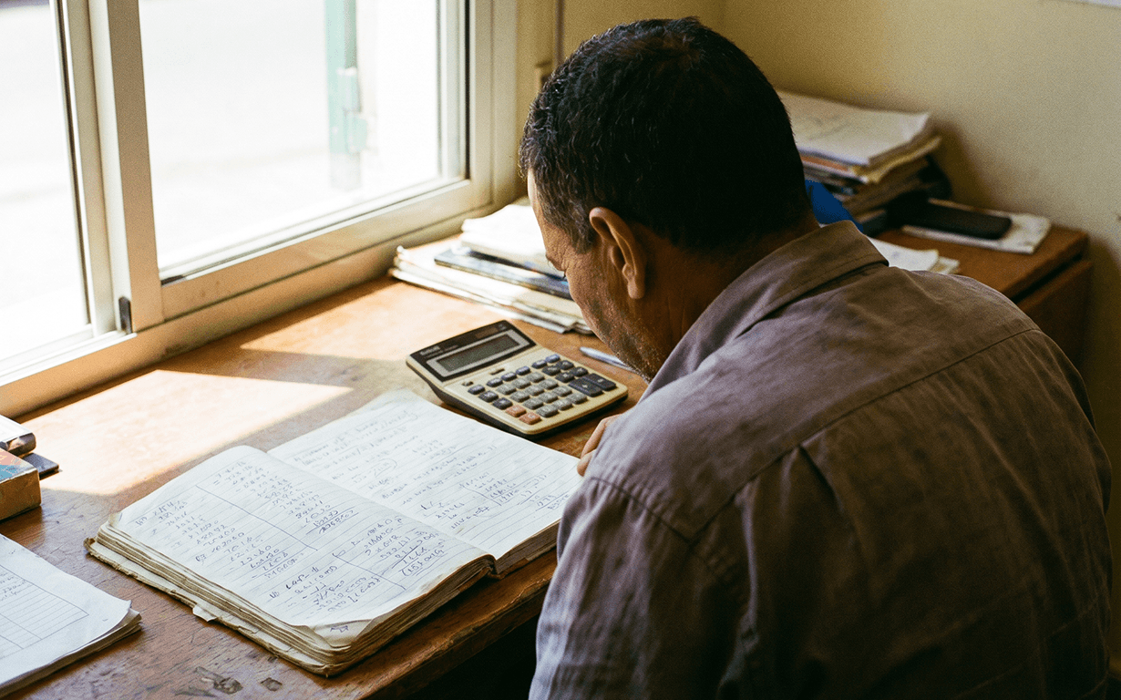 A man sits at a wooden desk by a window, reviewing handwritten notes in a ledger. A calculator and stacks of papers hint at his money sending routine as sunlight streams through the window.